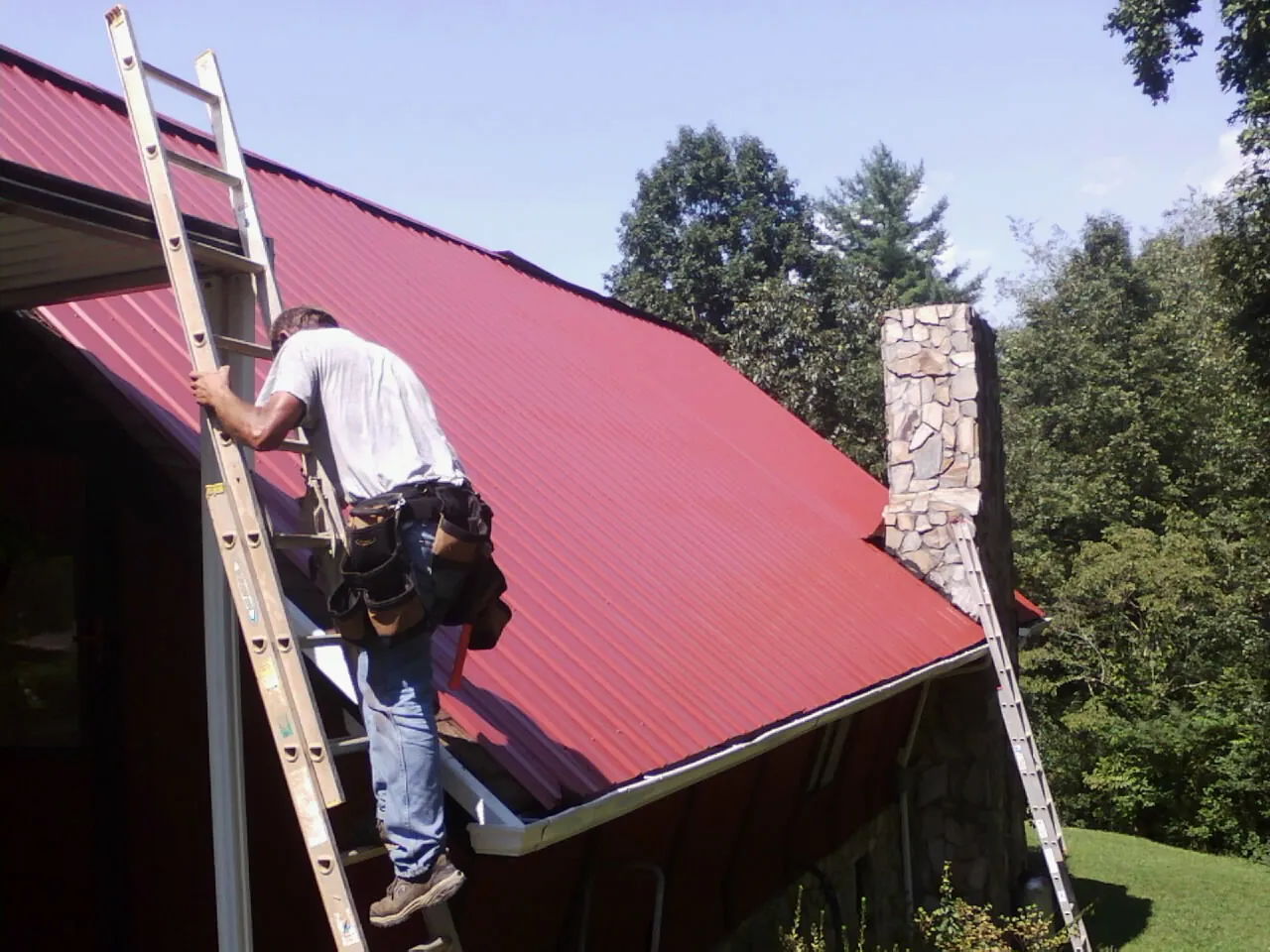 Roofer on a ladder beside a red metal roof and stone chimney — Soffit & Fascia Repair by Roofing Brothers in Wessington Springs